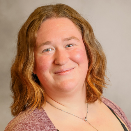 Professional headshot of a smiling person with shoulder-length wavy light brown hair wearing a maroon cardigan over a black top and a delicate silver necklace. The person is facing the camera against a soft gray studio background with warm, even lighting, creating a friendly, approachable, and professional appearance.
