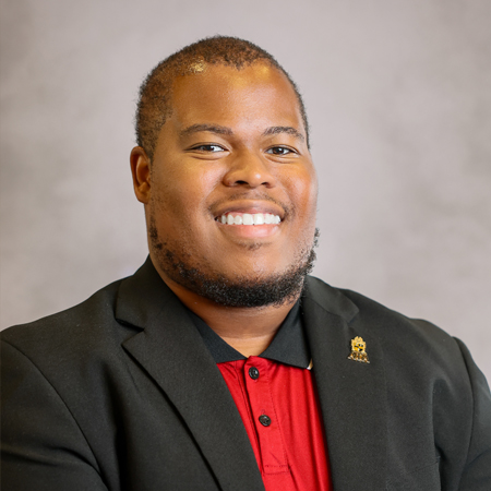 Professional headshot of a smiling person with closely cropped hair and a neatly trimmed beard wearing a black blazer over a red collared polo shirt. The person is facing the camera and is wearing a small lapel pin on the blazer. The photo is set against a soft gray studio background with warm, even lighting, creating a friendly, approachable, and professional appearance.