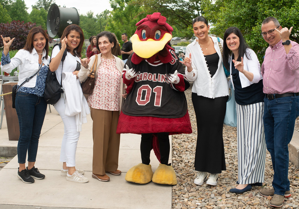 A student smiling while posing with Cocky along with family and friends at an outdoor event.