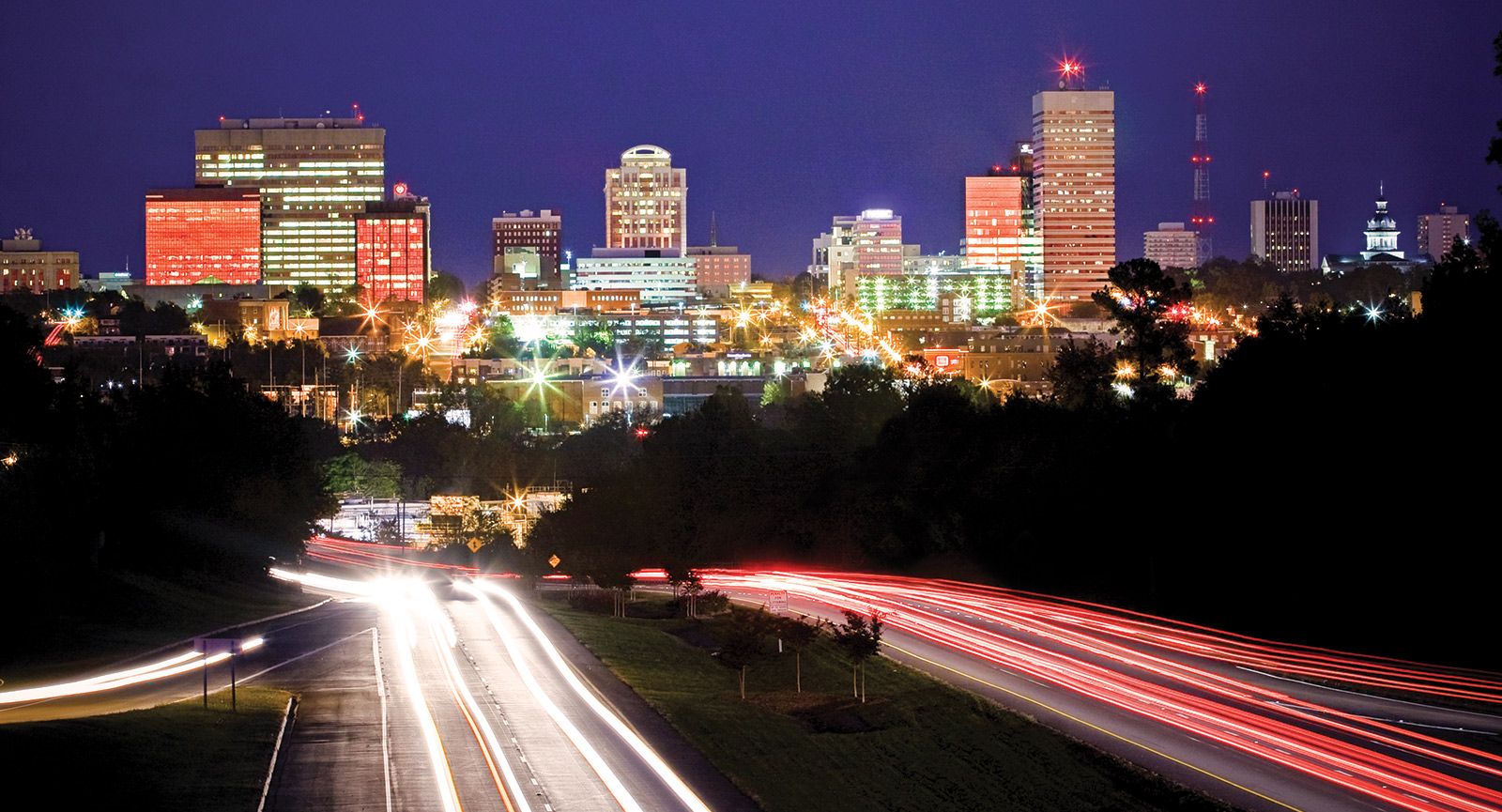 Car taillights streaming down the highway leading into the Columbia downtown skyline light up at night with glowing windows in the buildings. 