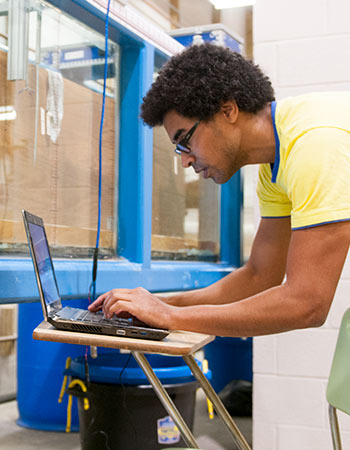 Student leans over a desk to type on a laptop in a lab setting.