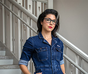 a woman with dark hair stands on a white stairwell with a jean jumpsuit on