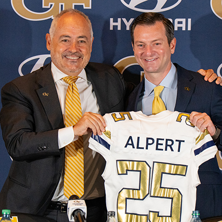 Two men in suits smile while holding a Georgia Tech football jersey with the name “Alpert” and number 25 during an introductory press conference.