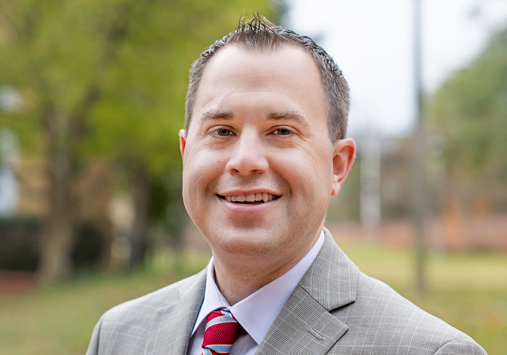 A headshot of Ryan Rucker wearing a suit and smiling outside Davis College on a cloudy day