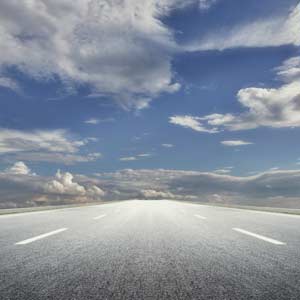 A view of a long, straight paved road with blue sky and clouds above.